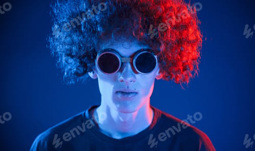 Preview: Portrait of young man with curly hair that is indoors illuminated by neon lighting. In retro eyewear
