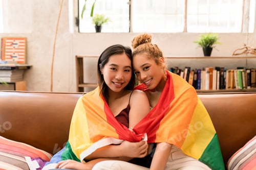 Preview: Two multiracial lesbian women in lgbt flag at home. Happy girlfriends couple at home
