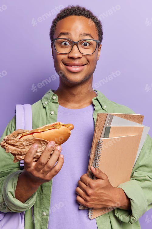 Preview: Vertical image of surprised dark skinned curly man holds hot dog and notepads eats during break