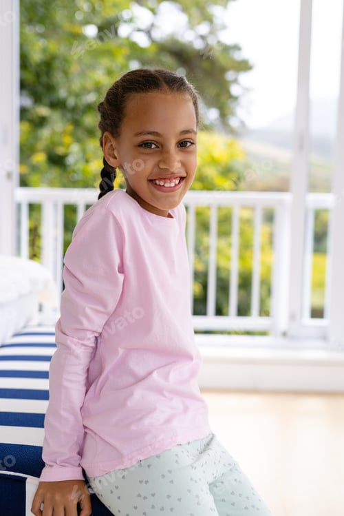 Preview: Portrait of happy biracial girl wearing pyjamas in bedroom in front of window with view of garden