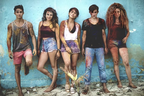 Preview: Five young people lined up against a wall covered in paint looking at camera.