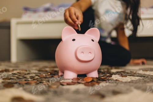Preview: diverse pre school girl at home in bedroom putting coins in pink piggy bank learning to save money