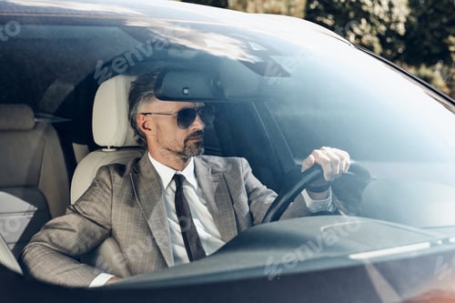 Preview: Confident mature man in formalwear and eyeglasses driving a car