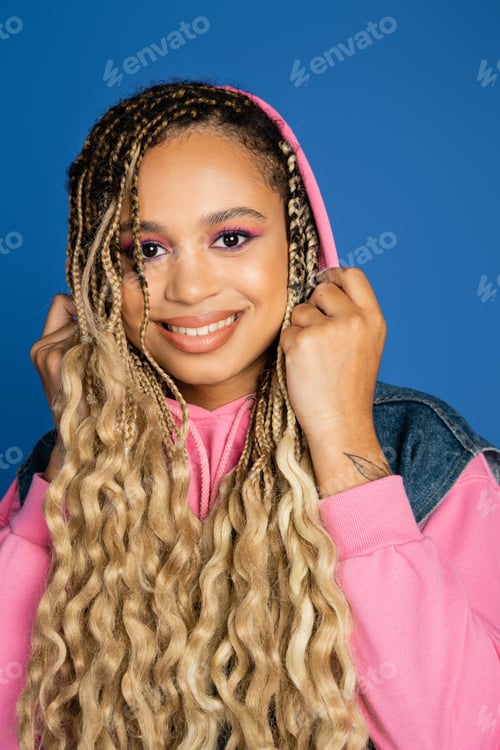 Preview: positive dark skinned woman with hood on head looking at camera and smiling on blue background