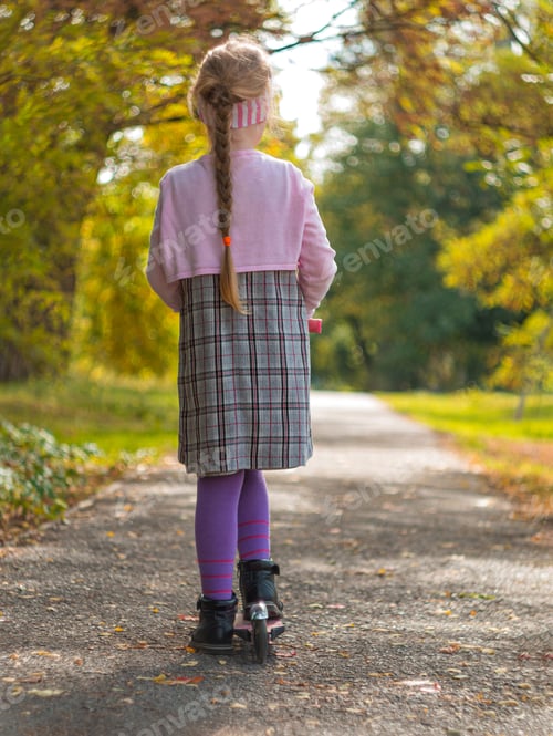 Preview: Child riding on kickscooter. Blonde girl in dress on scooter on road Autumn active lifestyle leisure