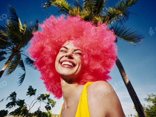 Preview: Bright pink afro woman, yellow sun hat, standing in front of palm trees