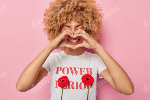 Preview: Positive curly haired woman makes heart gesture over mouth smiles toothily dressed in casual t shirt