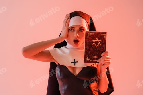Preview: shocked nun in sexy dress touching head while holding jewish bible isolated on pink