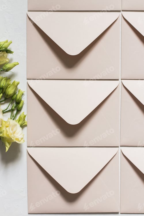 Preview: Vertical top view of cream envelopes on white surface next to flower