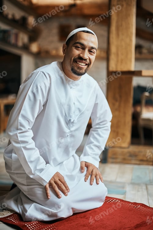 Preview: Happy Muslim on kneeling on a prayer mat at home and looking at camera.