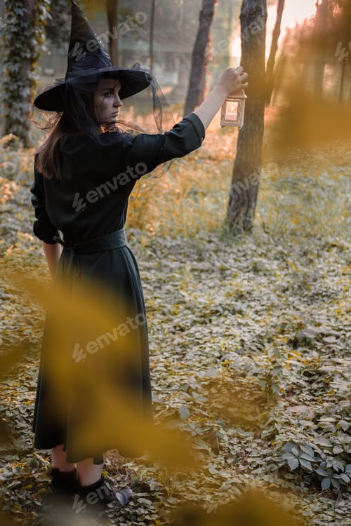 Preview: Young woman in dark dress and witch's hat holds lantern with candles and illuminates the forest.