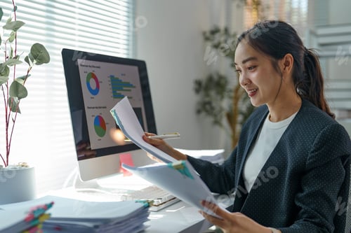 Preview: Young professional woman reviewing paperwork and comparing with charts displayed on computer screen,