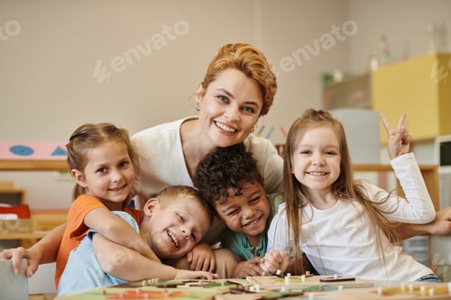 Preview: positive teacher hugging interracial kids and looking at camera during lesson in montessori school