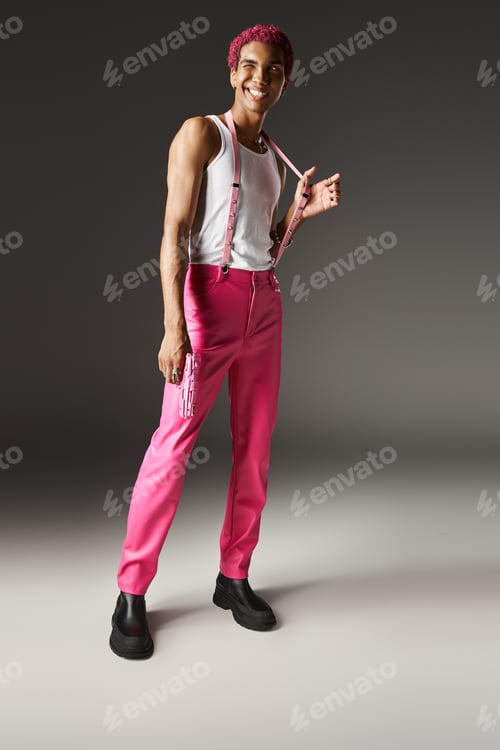 Preview: cheerful young man in pink attire winking and smiling holding pink toy gun on grey backdrop