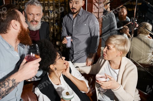 Preview: bearded men talking near amazed women, multiethnic colleagues with cocktails relaxing in bar