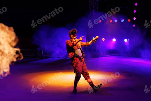Preview: Clown with a funny hairstyle entertains guests in the circus arena in light of spotlights and smoke