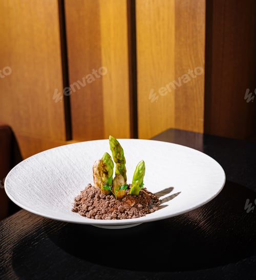 Preview: Green asparagus dish served on white plate with brown crumble on black table