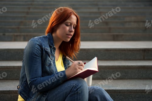 Preview: Young lady focused as she writes down notes in a journal with a pencil while seated on stone steps