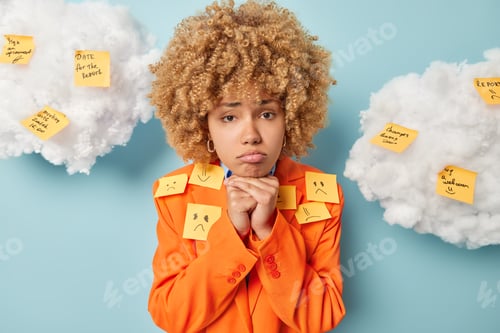 Preview: Upset doleful curly haired woman keeps hands under chin purses lips has offended expression has