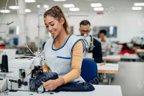 Preview: Happy female worker sewing while working at production line in clothing factory.
