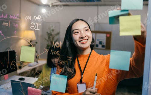 Preview: Young manager is smiling while organizing tasks for the day, writing on sticky notes and attaching t