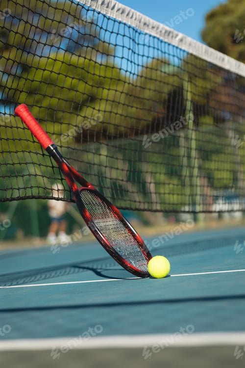 Preview: Surface level view of ball by racket leaning on tennis net at court during sunny day