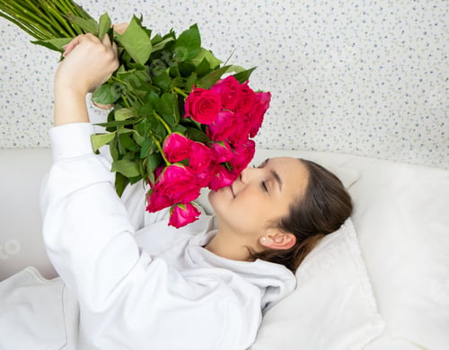 Vorschau: Ein junges und glückliches Mädchen liegt auf dem Bett und riecht mit geschlossenen Augen Rosen