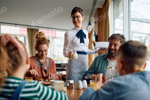 Preview: Happy waitress serving food and milk to a family who is eating breakfast in a hotel.