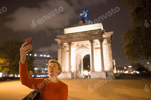 Preview: Mature female tourist taking smartphone selfie of Wellington Arch at night, London, UK