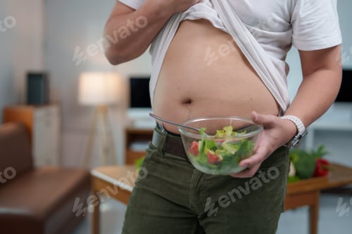 Preview: Overweight man lifting shirt showing belly holding bowl of salad