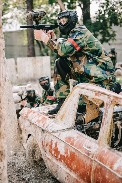 Preview: male paintball player aiming by marker gun and sitting on broken car while his team standing behind