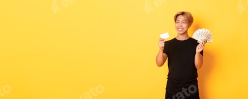 Preview: Portrait of satisfied smiling, handsome asian guy looking pleased at credit card while also holding