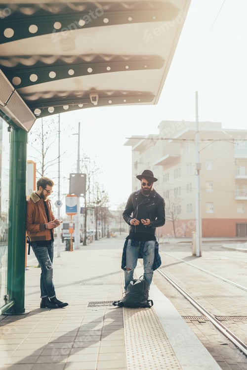 Preview: Two young male hipsters looking at smartphones while waiting at city tram station