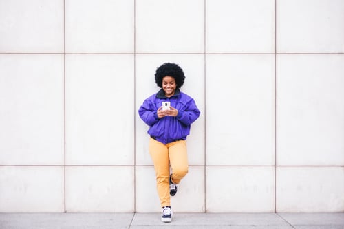 Preview: young latin afro woman standing on white wall using phone and smiling