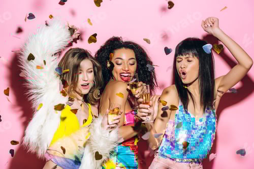 Preview: excited fashionable multicultural girls dancing with glasses of champagne on pink with confetti