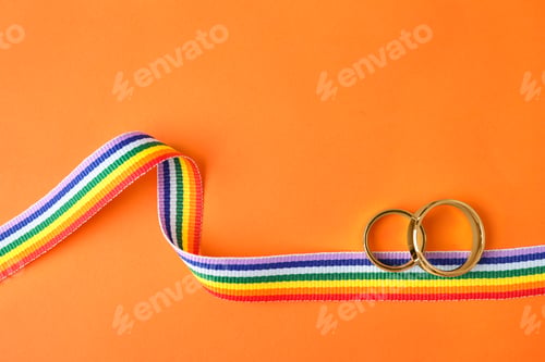 Preview: Wedding rings and rainbow ribbon on color background, top view. Gay symbol