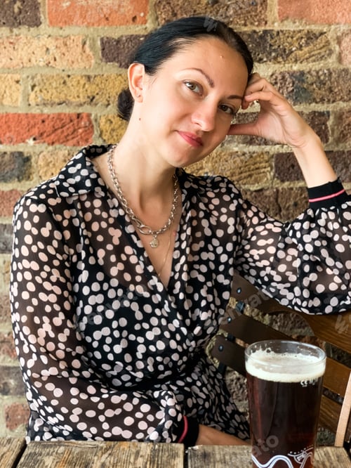 Preview: Young beautiful woman in old traditional pub drinking dark stout beer, wearing polka dot silk dress