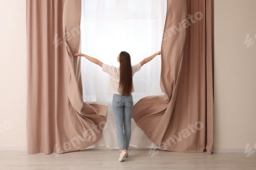 Preview: Woman opening stylish curtains at home, back view