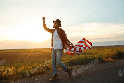 Preview: 4th of July. American Flag. Traveler with flag of America. Man in hat, backpack, a shirt and jeans.
