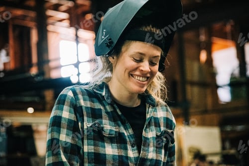 Preview: Bashful tradeswoman has her welding helmet lifted up as she smiles in her workshop feeling happy