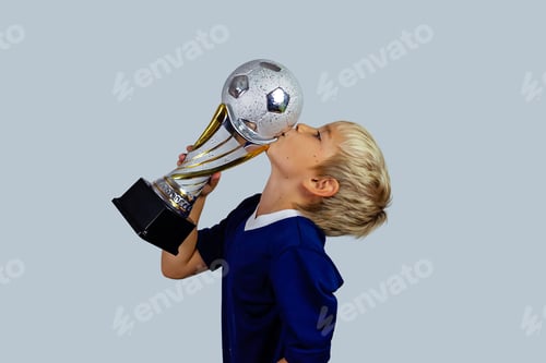 Preview: Young soccer player in blue jersey holds winners cup after the goal, isolated at light background