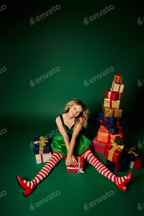 Preview: smiley woman in green dress and striped stockings sitting on floor with gifts, new year elf concept