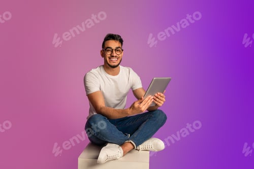 Preview: Full length of young Arab man using digital tablet, checking new app in neon light