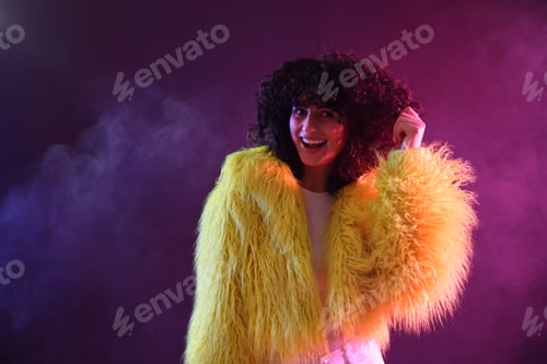 Preview: Beautiful young woman in yellow fur coat on color background in neon lights and smoke