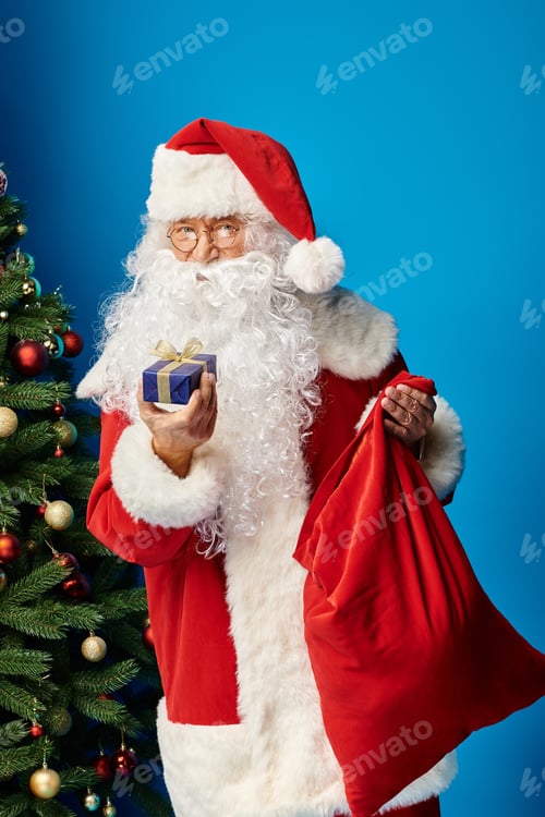 Preview: Santa Claus with beard and eyeglasses in red outfit holding sack bag and present near Christmas tree