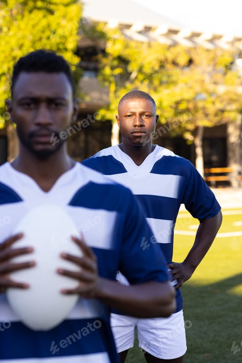Preview: Two African American young male athletes are holding a rugby ball on field outdoors, looking focused