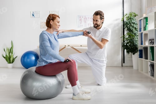 Preview: smiling physiotherapist helping redhead woman sitting on fitness ball and working out with dumbbells