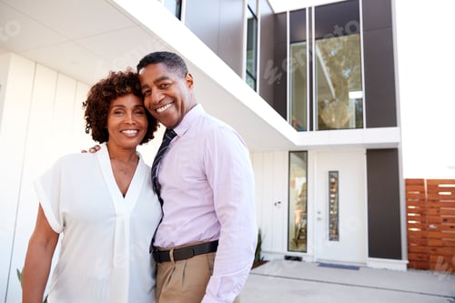 Preview: Middle aged black couple stand outside looking to camera in front of their modern home, close up