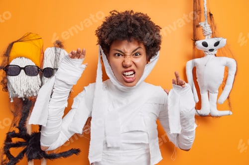 Preview: Annoyed young African American woman dressed like mummy clenches teeth raises arms celebrates hallow