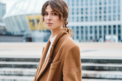 Preview: Young stylish woman in coat confidently looking in camera on city street